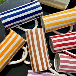 Peut inclure: Plusieurs mugs en céramique avec un motif rayé de différentes couleurs, dont le bleu, l'orange, le rouge et le marron. Chaque mug a une anse blanche et un intérieur blanc. Les mugs sont disposés sur une surface sombre, certains étant partiellement visibles.