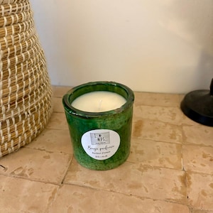 Peut inclure: Un bougeoir en céramique verte avec une bougie blanche à l'intérieur. Le bougeoir a une surface texturée et une étiquette avec du texte. La bougie est sur une surface carrelée, avec un panier tressé en arrière-plan.