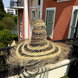 Peut inclure: Un abat-jour en paille tressée, de grande taille, avec un large bord et un sommet conique. L'abat-jour de couleur naturelle est suspendu à l'extérieur, projetant une ombre sur un rebord en béton. Le design présente des cercles concentriques de paille tressée.