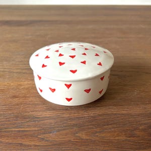 Peut inclure: Une petite boîte à bijoux en céramique blanche avec un couvercle. La boîte est décorée d'un motif de cœurs rouges.