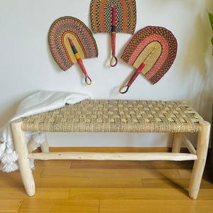 May include: Three woven wall fans in red, orange, yellow, black, and green colours hanging on a white wall above a natural woven bench with wooden legs.