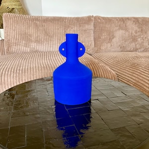 Peut inclure: Un vase en céramique bleu vif de forme originale, avec deux poignées circulaires sur les côtés. Le vase est posé sur une table sombre et brillante, carrelée, reflétant sa couleur. Le fond comprend un canapé en velours côtelé.