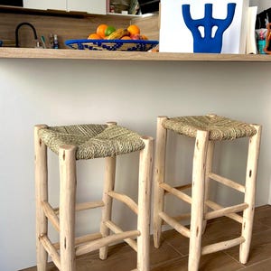 Peut inclure: Deux tabourets de bar rustiques en bois avec des assises tressées. Les tabourets ont des pieds en bois de couleur claire et une assise tressée naturelle. Les tabourets sont placés devant un comptoir de cuisine avec un objet décoratif bleu et un bol de fruits.