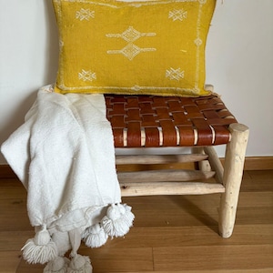 Peut inclure: Un coussin décoratif jaune moutarde avec broderie géométrique blanche repose sur un banc en cuir tressé. Une couverture blanche avec une garniture à gros pompons est drapée sur le banc. Le banc a un cadre en bois clair.