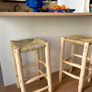 May include: Two wooden bar stools with woven straw seats. The stools have a natural wood finish and a simple, sturdy design. The seats are square and appear comfortable. The stools are positioned in front of a light-coloured countertop.