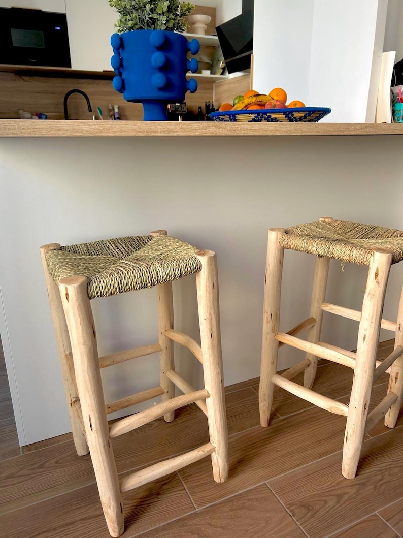 May include: Two wooden bar stools with woven straw seats. The stools have a natural wood finish and a simple, sturdy design. The seats are square and appear comfortable. The stools are positioned in front of a light-coloured countertop.