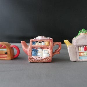 May include: Three decorative ceramic teapots. The first is brown with a red handle and spout, resembling a radio. The second is pink with a shelf design. The third is grey with a yellow handle.
