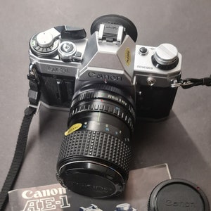 Könnte beinhalten: Eine silberne Canon AE-1 Kamera mit einem schwarzen Canon Objektiv. Die Kamera liegt auf einer schwarz-weißen Bedienungsanleitung mit dem Text "Canon AE-1" und "Canon International Service Network" darauf.