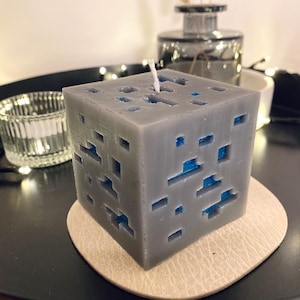 Könnte beinhalten: Eine graue, würfelförmige Kerze mit blauen Akzenten, die einem pixeligen Design ähnelt. Die Kerze steht auf einem beigen Untersetzer, im Hintergrund befinden sich ein Glaskerzenhalter und eine Vase.