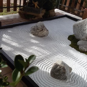 Könnte beinhalten: Ein Miniatur-Zen-Garten mit weißem Sand, der in kreisförmigen Mustern geharkt wurde. Mehrere graue Steine unterschiedlicher Größe sind auf dem Sand platziert, einige ruhen auf einem grünen Moospolster. Der Garten ist in einen schwarzen Rahmen eingefasst.