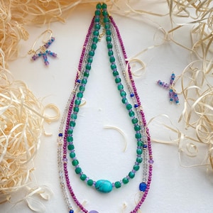 May include: A layered necklace with green, pink and grey beads, featuring a turquoise gemstone and a clear crystal. Matching earrings with purple and blue beads are also included.