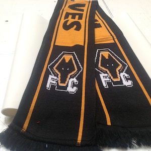 May include: Black and orange knitted scarf with a white wolf head logo and the text "FC" on it. The scarf has a fringe on both ends.