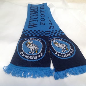 May include: Blue and black knitted scarf featuring the Wycombe Wanderers Football Club logo. The logo depicts a white bird with a yellow beak and a yellow object in its beak. The text "Wycombe Wanderers" is written in white on a blue background.