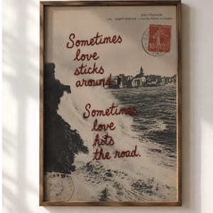 Könnte beinhalten: Ein gerahmter Kunstdruck mit einer schwarz-weißen Vintage-Küstenlandschaft mit brechenden Wellen und einer Stadt im Hintergrund. Roter, gestickter Text lautet: Sometimes love sticks around. Sometimes love hits the road. Inklusive roter Briefmarke.