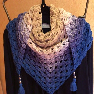 Könnte beinhalten: Ein gehäkeltes Tuch mit einem Farbverlauf von Blau, Lila und Beige. Das Tuch hat einen gewellten Rand und ist mit silbernen Perlen und Quasten verziert.