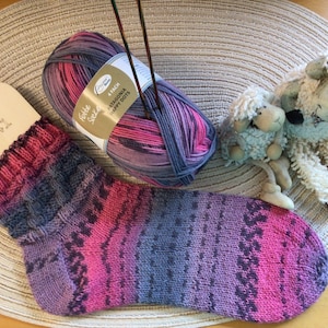 Könnte beinhalten: Ein Paar handgestrickte Socken in Pink und Lila mit einem Wollknäuel und Stricknadeln. Das Wollknäuel ist mit "Felle Socke" und "Niagara Happy Dots" beschriftet.