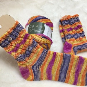 Könnte beinhalten: Ein Paar handgestrickte Socken in einem bunten Streifenmuster aus Orange, Gelb, Lila und Rosa. Die Socken sind aus einem weichen, flauschigen Garn gefertigt und werden neben einem Knäuel Garn gezeigt, das mit "Fluffy Silk 4 fach Silk Socks" beschriftet ist.
