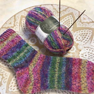 Könnte beinhalten: Eine handgestrickte Socke in leuchtenden Regenbogenfarben, zusammen mit einem Knäuel passender Wolle und Stricknadeln. Das Garnetikett lautet "Flotte Socke Alpaka-Merino". Die Socke und das Garn sind eine Mischung aus Rosa, Lila, Grün und Gelb.