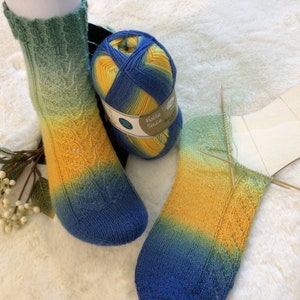 Könnte beinhalten: Zwei Paar handgestrickte Socken in einem Farbverlauf von Grün, Gelb und Blau. Das Garn ist ein mehrfarbiger Strang in Blau, Gelb und Grün. Die Socken werden mit Holzstricknadeln gearbeitet.