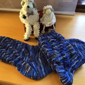 Könnte beinhalten: Ein Paar gestrickte Socken in Blau, Grau und Weiß. Die Socken liegen auf einer Holzoberfläche. Im Hintergrund befinden sich zwei kleine weiße Schaffiguren.