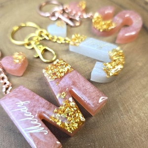 May include: A collection of handmade resin keychains and charms. The keychains are in the shape of letters and hearts, with gold flakes embedded in the resin. The letters are in a pink hue, with the name "Melody" written on one of them.