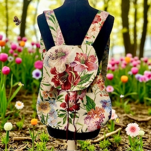 Op de afbeelding: Bloemen tapestry rugzak met verstelbare bandjes. De rugzak heeft een crèmekleurige achtergrond met een levendig patroon van rode, roze en paarse bloemen en groene bladeren. De rugzak wordt getoond op een zwarte mannequin.