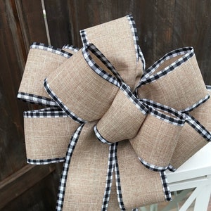 May include: A decorative bow made of tan burlap ribbon with a black and white checkered trim. The bow is placed on top of a white lantern with glass panels. The lantern has a classic design, perfect for home decor.