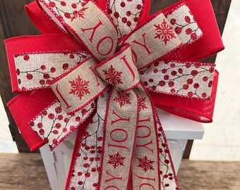 May include: A red and beige Christmas bow with the word "JOY" repeated on the tails. The bow is decorated with red berries and snowflakes.