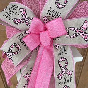 May include: Pink and beige ribbon bow with a leopard print and pink ribbon design. The ribbon has the words "Be Brave", "Have Hope", "Faith", and "Be Strong" printed on it.