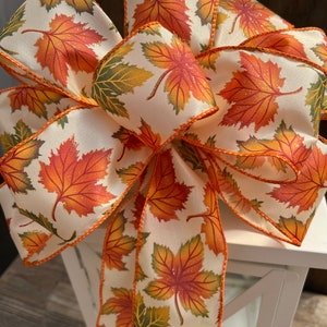 May include: A decorative bow featuring a cream-colored ribbon adorned with colorful autumn leaves in shades of orange, red, and green. The ribbon has an orange trim. The bow is likely used for seasonal decor, such as fall or Thanksgiving.