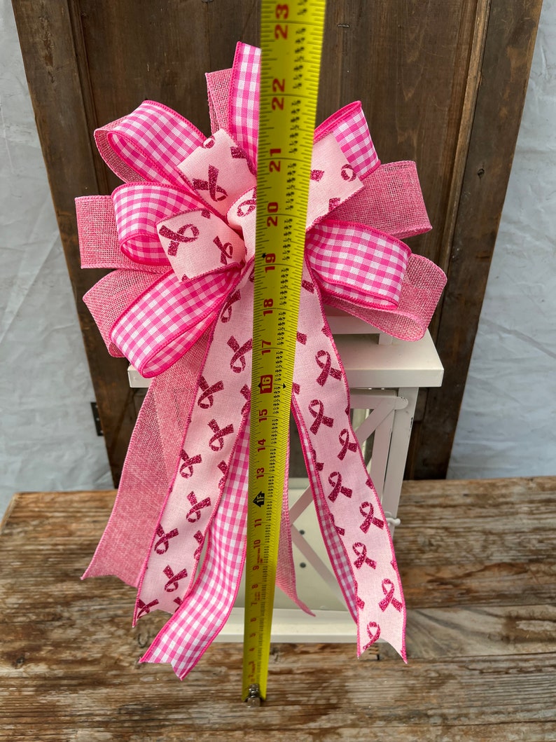 May include: A pink and white gingham ribbon bow with pink glitter ribbon accents. The bow is tied around a white lantern. The bow is approximately 18 inches tall.