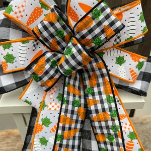 May include: A black and white checkered ribbon with a white ribbon with orange carrots and green leaves printed on it. The ribbons are tied together in a bow.