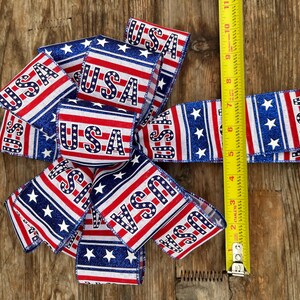 May include: A red, white, and blue ribbon with stars and the word "USA" printed on it. The ribbon is folded into a bow shape and is being measured with a tape measure.