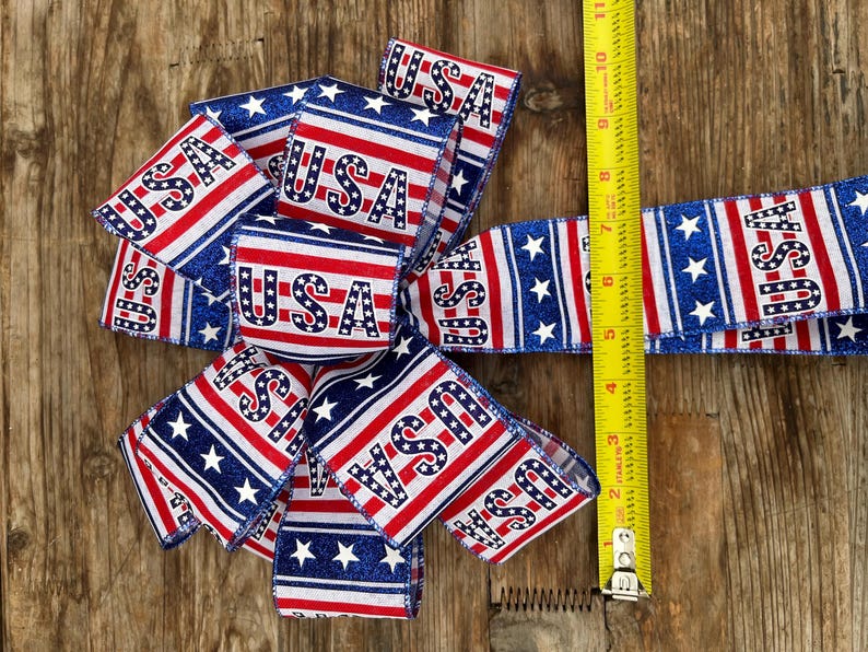 May include: A red, white, and blue ribbon with stars and the word "USA" printed on it. The ribbon is folded into a bow shape and is being measured with a tape measure.