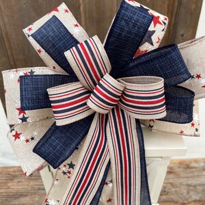 May include: A red, white, and blue striped ribbon bow with a navy blue and white star patterned fabric. The bow is tied around a white lantern.