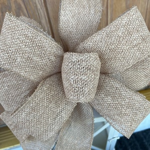 May include: A large, brown burlap bow with a woven texture. The bow is tied with a long tail of burlap ribbon.
