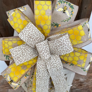 May include: A yellow and brown bow with a honeycomb pattern and bees. The bow is tied with a white and brown ribbon with a floral pattern.