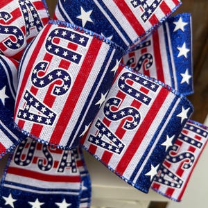 May include: A red, white, and blue ribbon bow with the word "USA" printed on it. The ribbon has a glittery blue stripe and white stars.