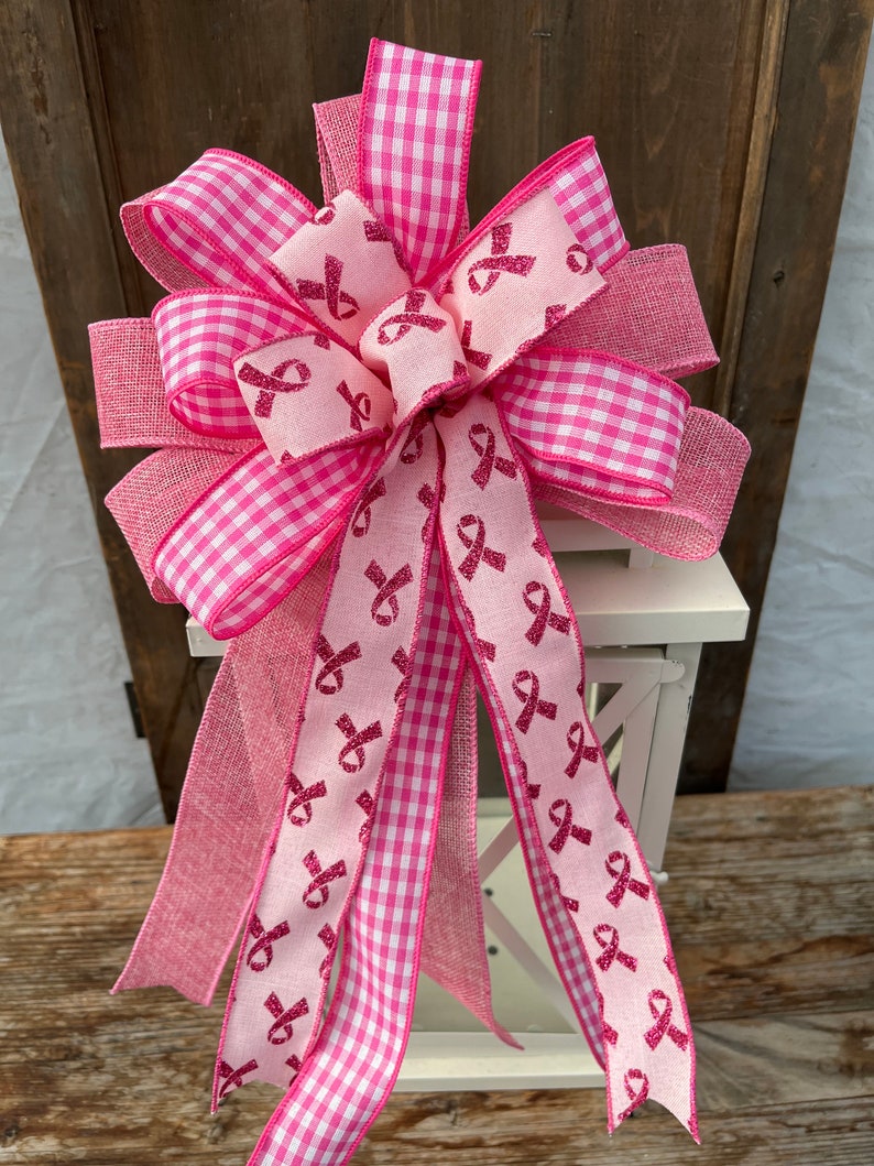 May include: A pink and white gingham ribbon bow with pink ribbon tails featuring a pink glitter ribbon with pink ribbon designs. The bow is tied around a white lantern.