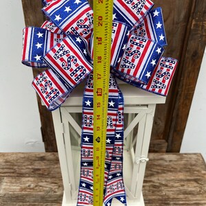 May include: A white lantern with a red, white, and blue ribbon bow. The ribbon has stars and the word "USA" printed on it. The ribbon is tied around the top of the lantern.