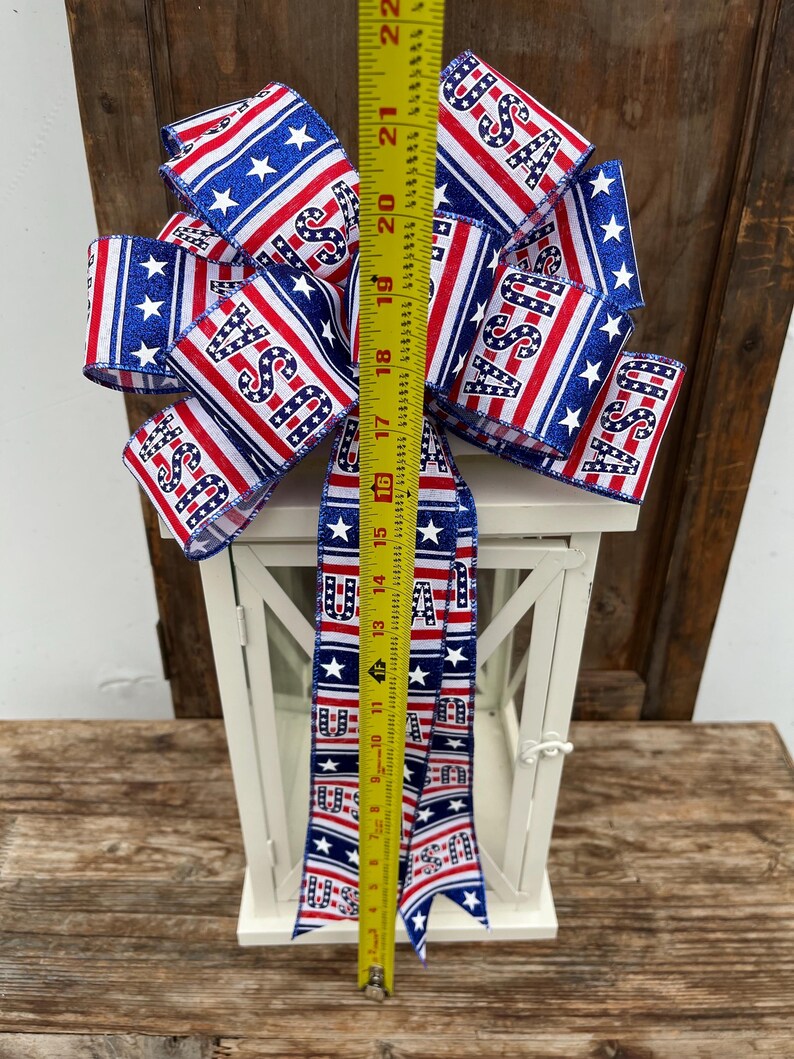 May include: A white lantern with a red, white, and blue ribbon bow. The ribbon has stars and the word "USA" printed on it. The ribbon is tied around the top of the lantern.