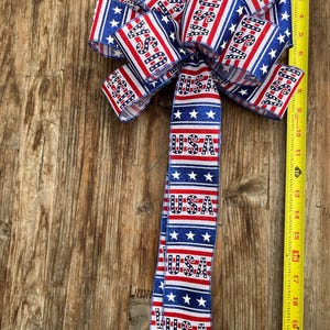 May include: A red, white, and blue bow with glitter and the word "USA" printed on it. The bow is tied with a ribbon that has the same pattern and is hanging from a wooden surface.