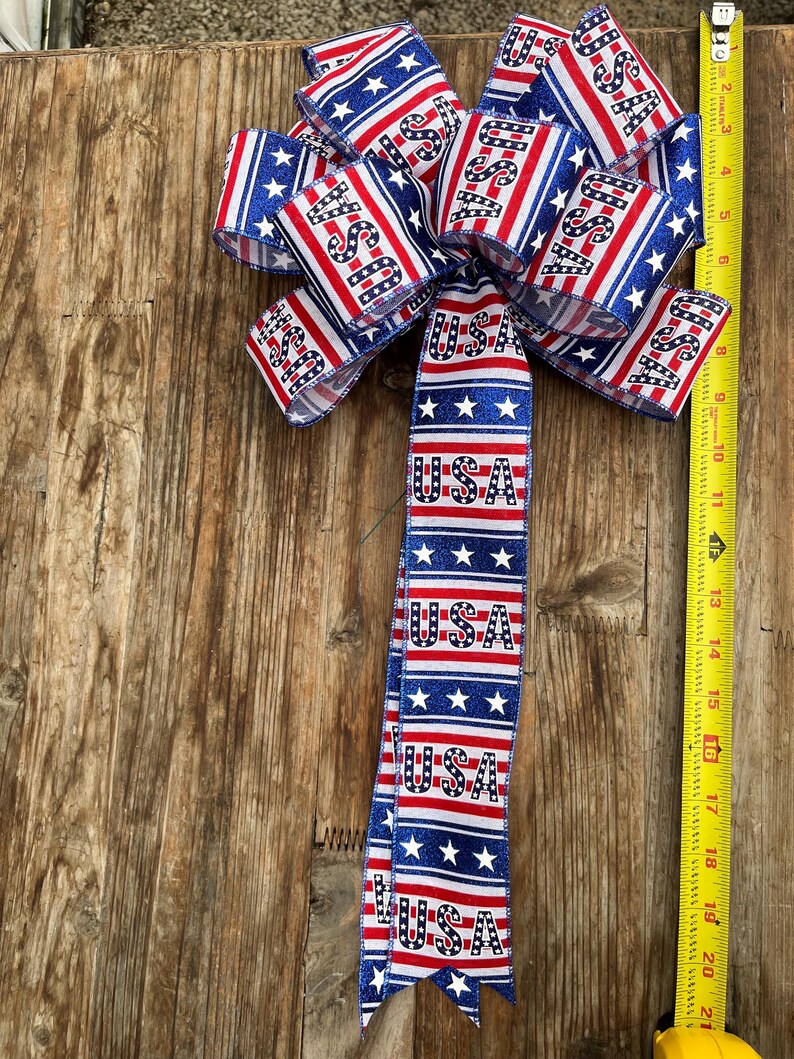 May include: A red, white, and blue bow with glitter and the word "USA" printed on it. The bow is tied with a ribbon that has the same pattern and is hanging from a wooden surface.