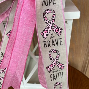 May include: A pink and beige ribbon with a pink leopard print ribbon and the words "have hope", "be brave", "have faith", and "be strong" printed on it.