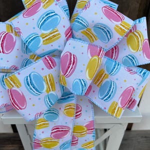 May include: A decorative bow featuring a white ribbon printed with colorful macarons in pink, yellow, and blue. The bow has a light blue stitched edge and is tied in a multi-loop style, perfect for gift wrapping or crafting.