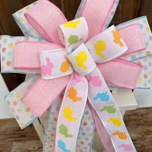 May include: A pink and blue polka dot ribbon with a white ribbon featuring pastel colored bunny silhouettes. The ribbons are tied into a bow.