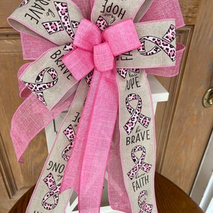 May include: Pink and brown ribbon bow with the words "be brave", "have hope", "be strong", and "have faith" printed on it. The bow is tied around a white lantern.