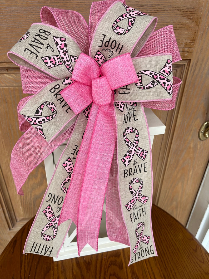 May include: Pink and brown ribbon bow with the words "be brave", "have hope", "be strong", and "have faith" printed on it. The bow is tied around a white lantern.