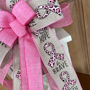 May include: A pink and beige ribbon bow with a pink leopard print ribbon. The ribbon has the words "have hope", "be brave", and "have faith" printed on it.