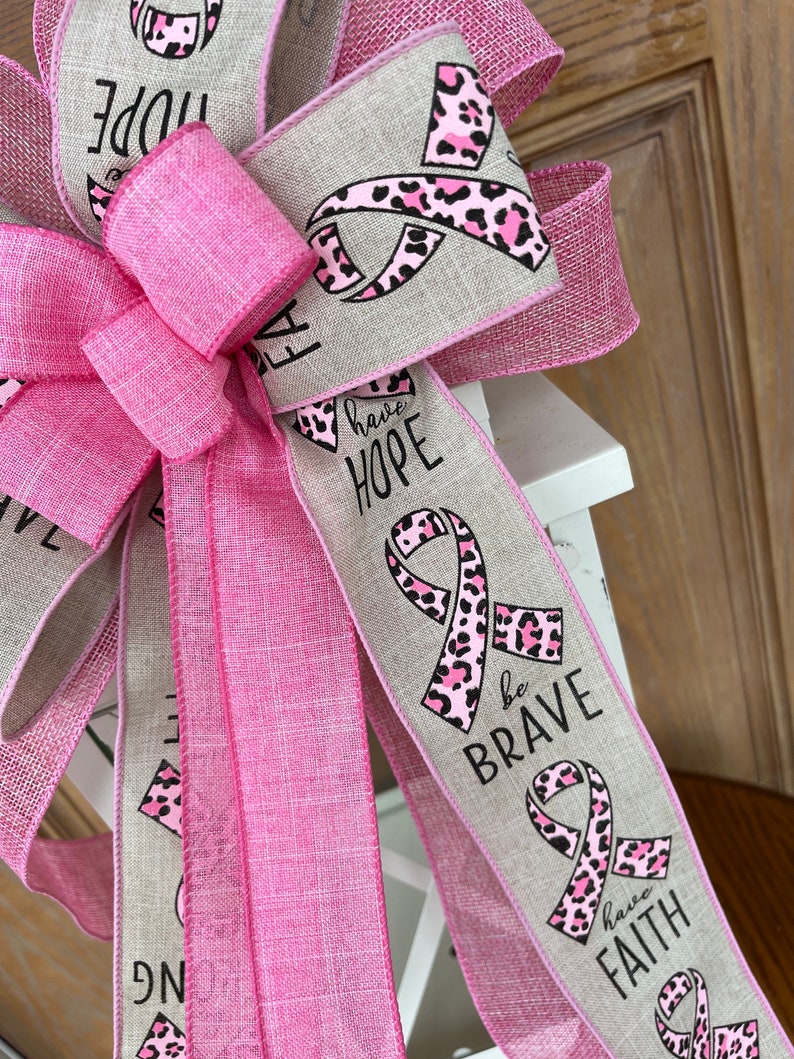 May include: A pink and beige ribbon bow with a pink leopard print ribbon. The ribbon has the words "have hope", "be brave", and "have faith" printed on it.
